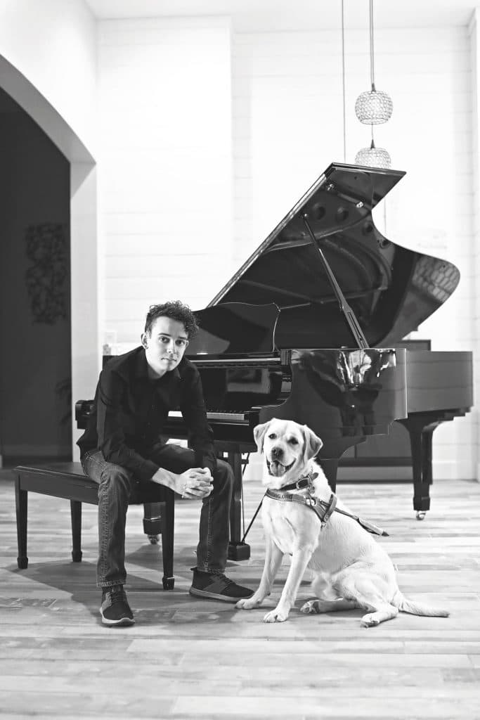 Brenden sits on a piano bench in front of an open black grand piano, hands clasped, looking at the camera. His light-coated guide dog, Diesel, sits beside him in a harness. Black-and-white photo.